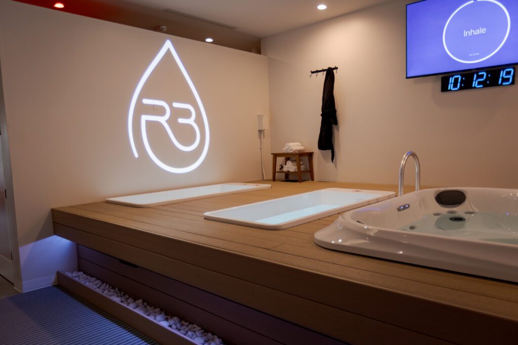 Treatment room for R3 wellness spa featuring a wooden deck with two cold plunge tubs, a hot tub, and a glowing R3 logo on the wall.