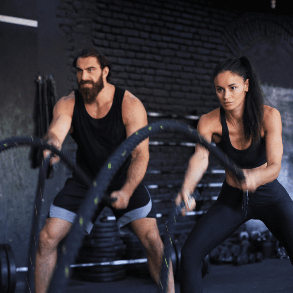 A man with a large beard and a woman in athletic wear performing high-intensity battle rope exercises in a gym.
