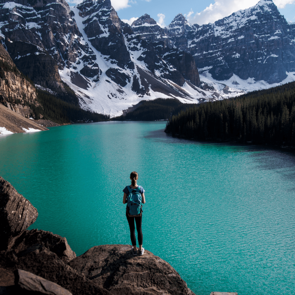 A woman with a backpack standing on a rocky ledge, looking out over the turquoise waters of Moraine Lake and the snow-capped Rocky Mountains.