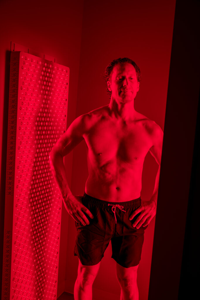 A shirtless, athletic man standing in front of a vertical red light therapy panel in a dark room, illuminated by a vibrant red glow.