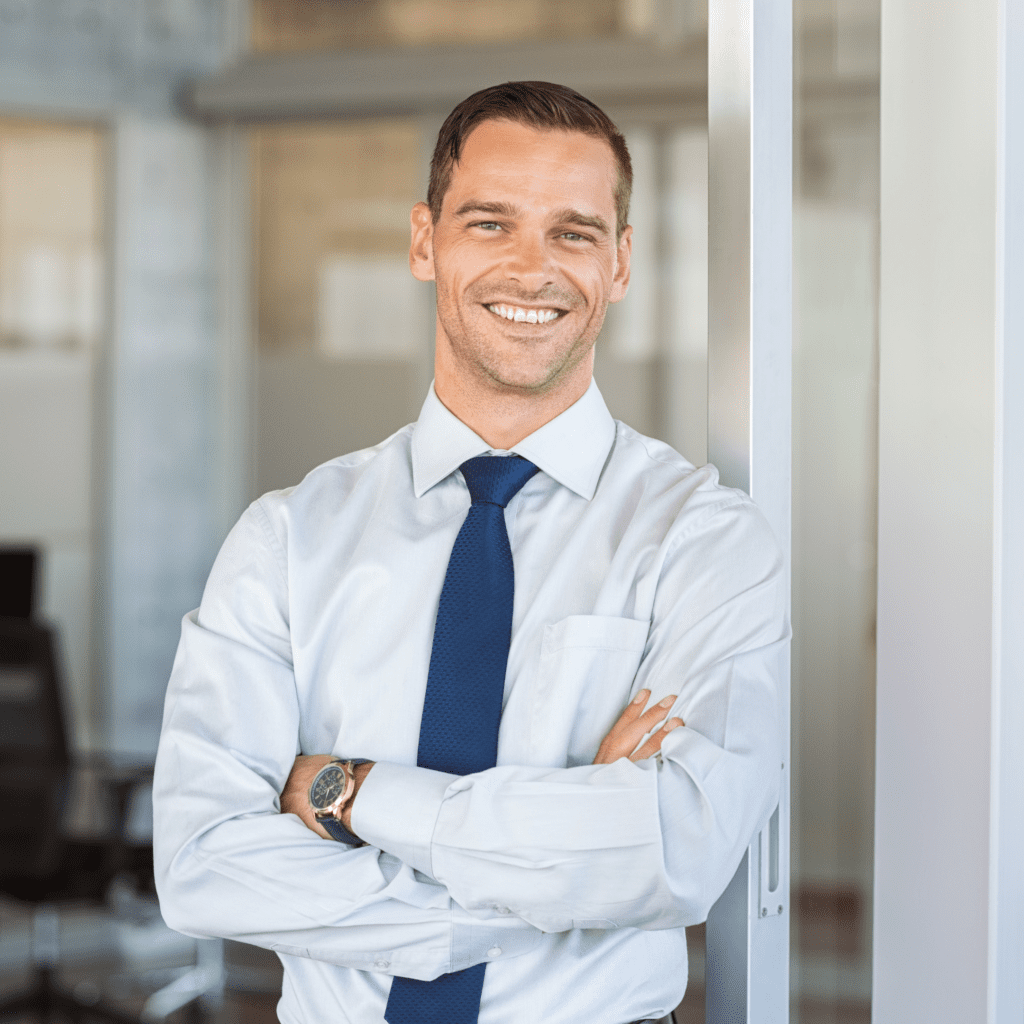 A smiling man in a white dress shirt and blue tie standing with his arms crossed and leaning against a white wall.