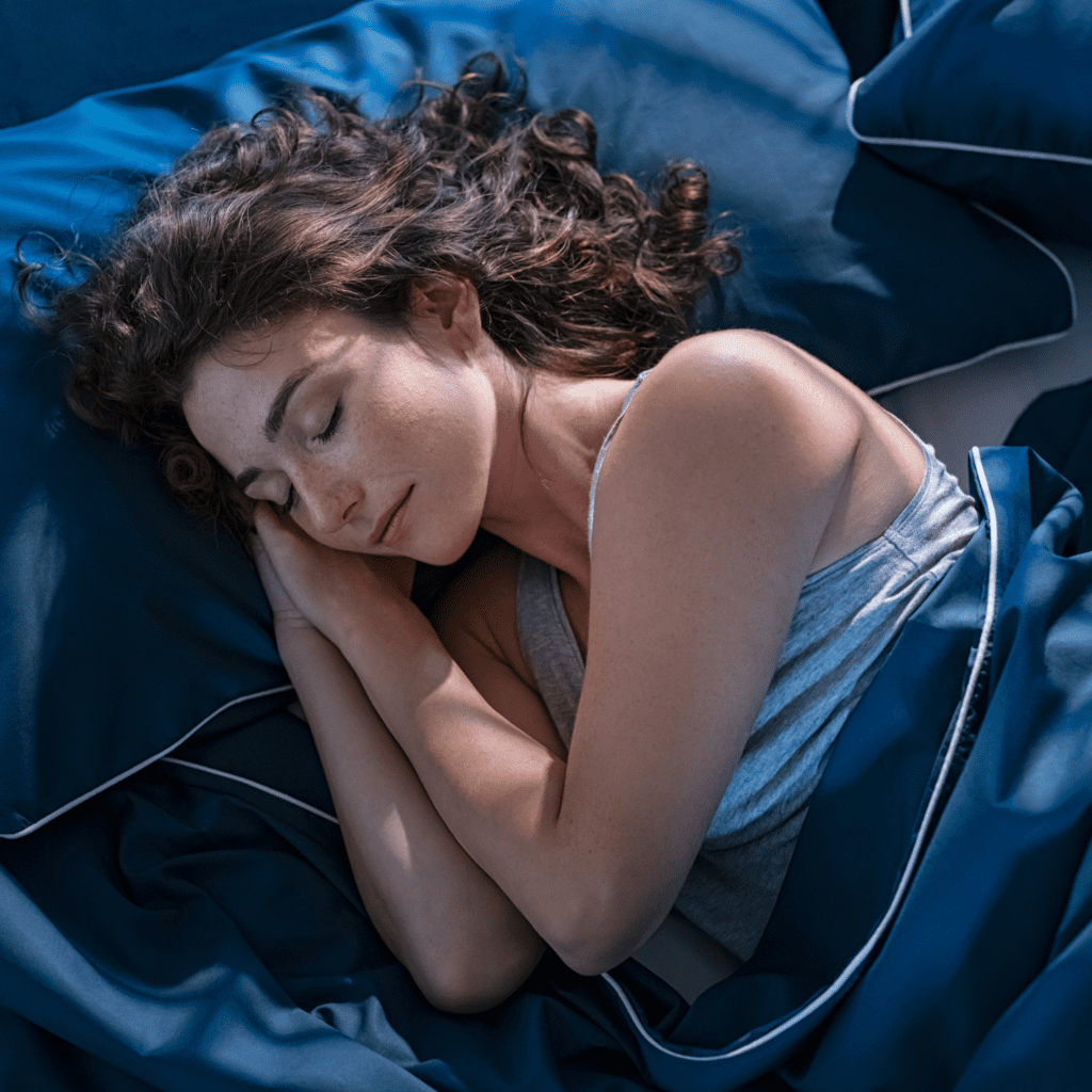 A woman with wavy brown hair sleeping peacefully on a pillow under a dark blue duvet.