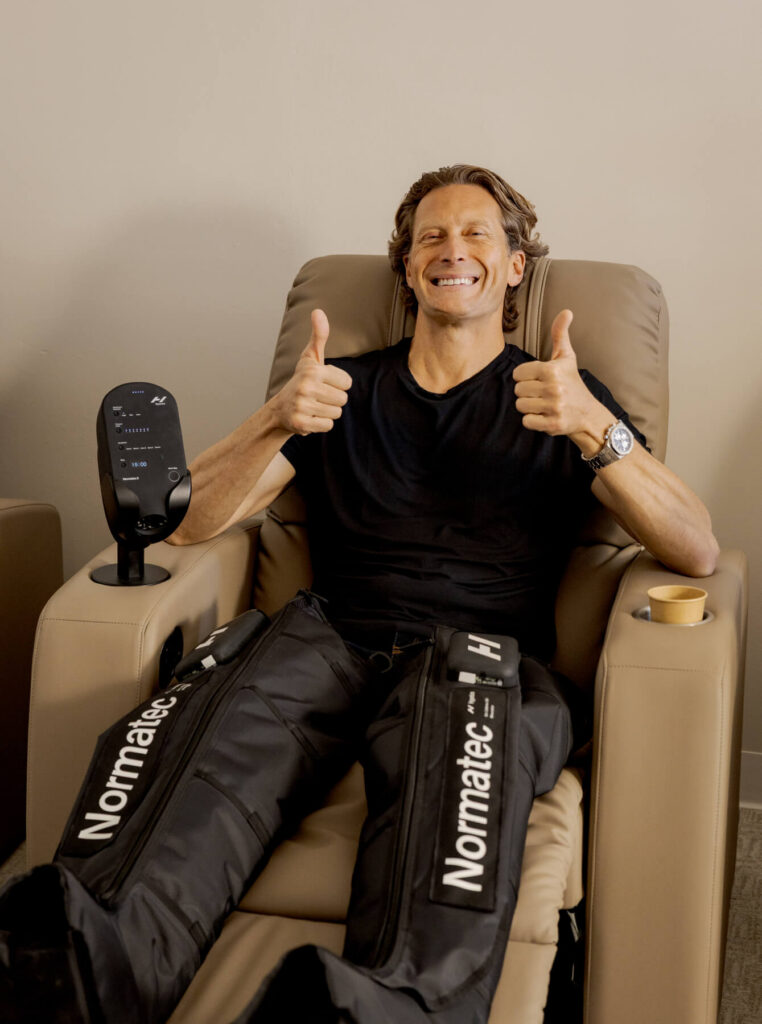 Man in a black tshirt sitting in a chair wearing Normatec compression legs with two thumbs up at R3