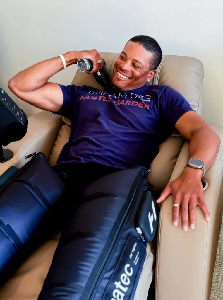 A smiling man in a blue t-shirt relaxes in a leather therapy chair while using a handheld massage device on his shoulder and wearing black Normatec compression boots.