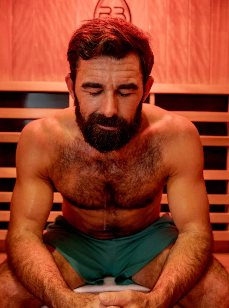 A shirtless man with a thick beard sitting in a wooden sauna, looking downward with a focused or meditative expression in the warm, red-toned light.