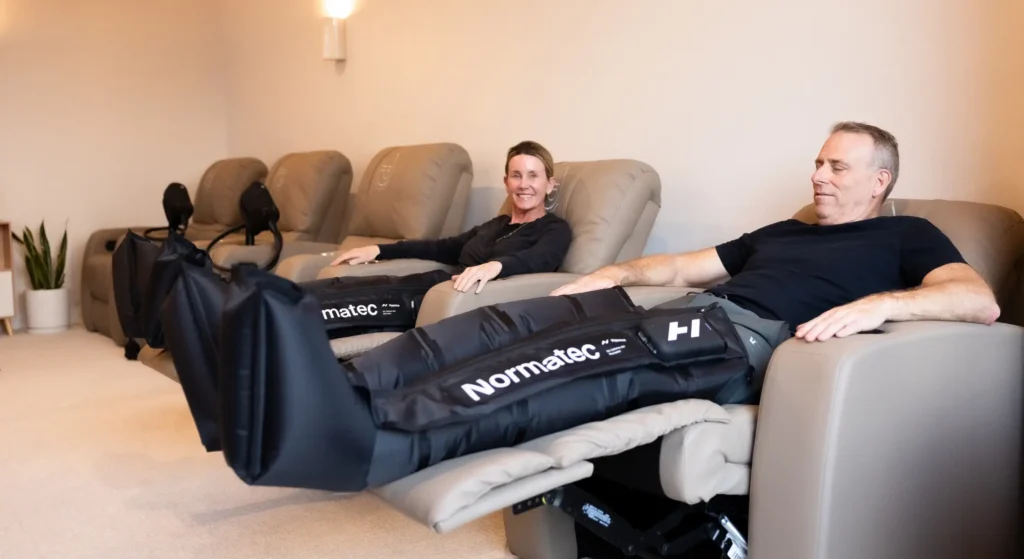 A man and a woman relaxing in leather treatment recliners while wearing black Normatec compression therapy boots on their legs.