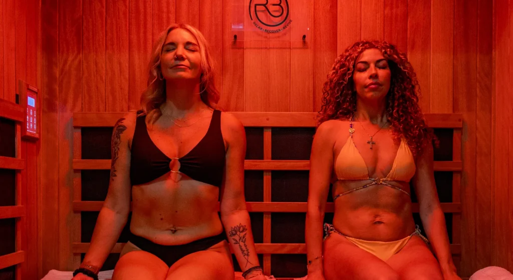 Two women in bikinis sitting on a wooden bench inside a sauna with red ambient lighting and the R3 logo on the back wall.