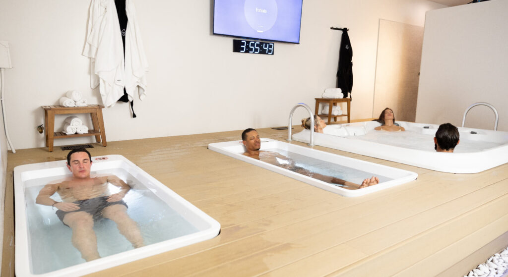 Multiple people relaxing in cold plunge tubs and a hot tub at a wellness spa with a guided breathing timer displayed on the wall